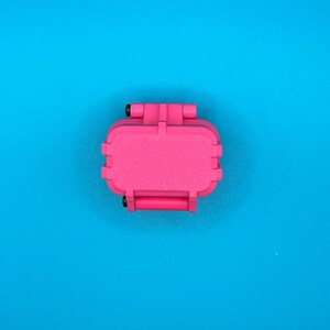 May include: A small, rectangular, hot pink storage case with a textured surface and a hinged lid. The case features two black circular hinge pins on the side, providing a secure closure mechanism for the compact, solid-colored plastic container.