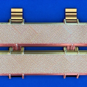 May include: Two rectangular, gold-colored, decorative tiles with a textured surface. The tiles are connected by a hinge and have gold-colored metal brackets on the top and bottom.