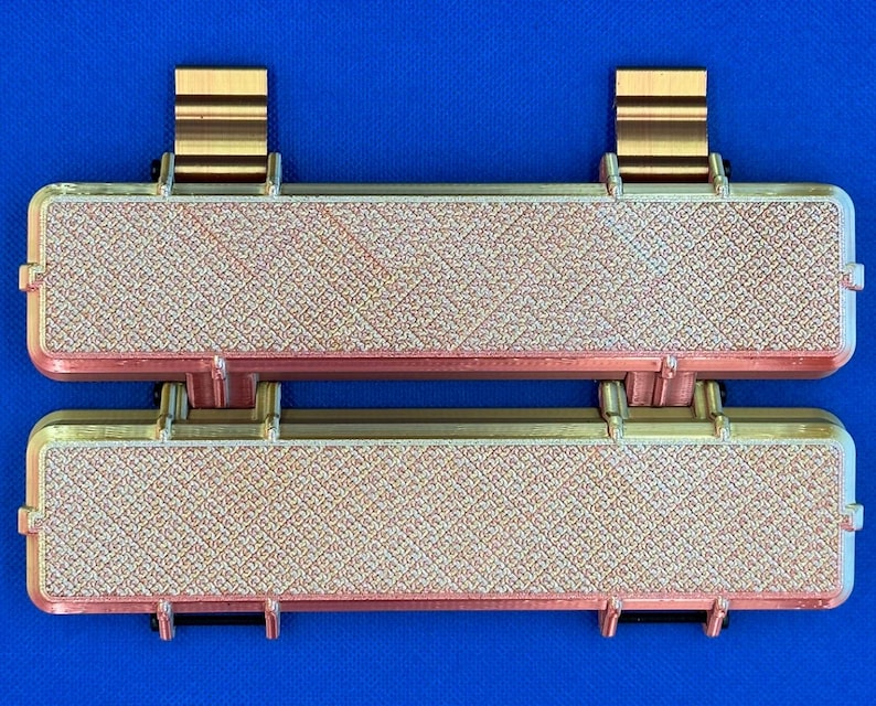 May include: Two rectangular, gold-colored, decorative tiles with a textured surface. The tiles are connected by a hinge and have gold-colored metal brackets on the top and bottom.
