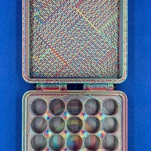 May include: A square, iridescent, multi-colored storage case with a hinged lid. The interior of the lid features a textured, cross-hatched pattern, while the base contains a grid of twenty circular, concave compartments with a spiral finish.