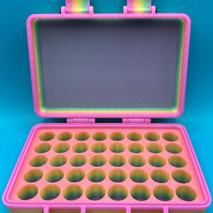 May include: A rectangular storage case with a hinged lid, featuring a multi-colored rainbow gradient finish. The interior base contains a grid of thirty-two circular compartments for small items, while the lid interior is flat and grey.