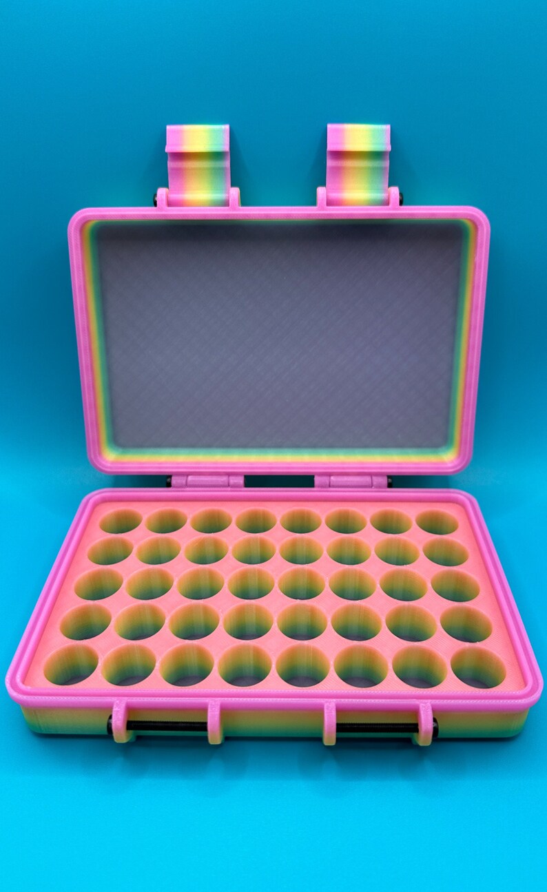 May include: A rectangular storage case with a hinged lid, featuring a multi-colored rainbow gradient finish. The interior base contains a grid of thirty-two circular compartments for small items, while the lid interior is flat and grey.