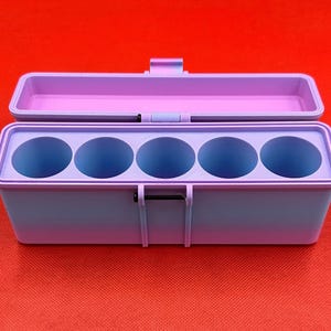 May include: A rectangular storage case with a hinged lid, featuring a gradient color scheme that transitions from light purple to blue. The interior contains five circular compartments arranged in a single row, designed for holding cylindrical items.