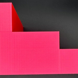 May include: A bright pink tiered display stand featuring four distinct levels of varying heights. The structure has a stepped design with a smooth, matte finish and a semi-circular cutout on the lowest level, providing a functional surface for organizing items.
