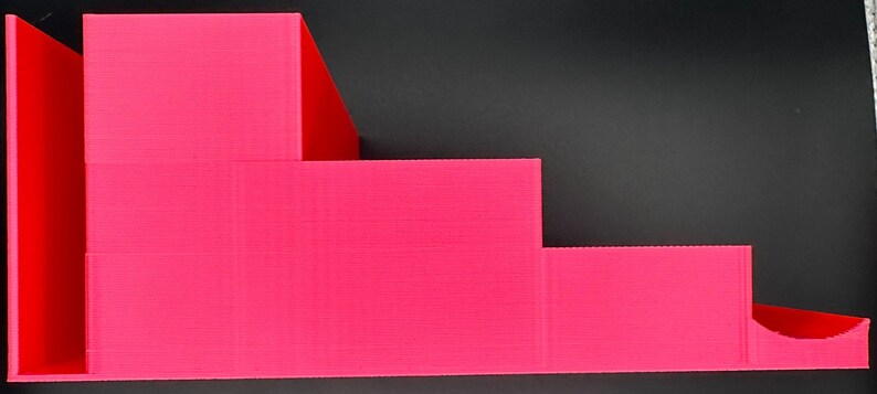 May include: A bright pink tiered display stand featuring four distinct levels of varying heights. The structure has a stepped design with a smooth, matte finish and a semi-circular cutout on the lowest level, providing a functional surface for organizing items.