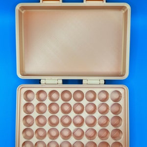 May include: A rectangular, metallic gold storage case with an open lid. The interior base features a grid of forty circular indentations arranged in five rows of eight, designed for holding small items. The case has two hinges and two latches.