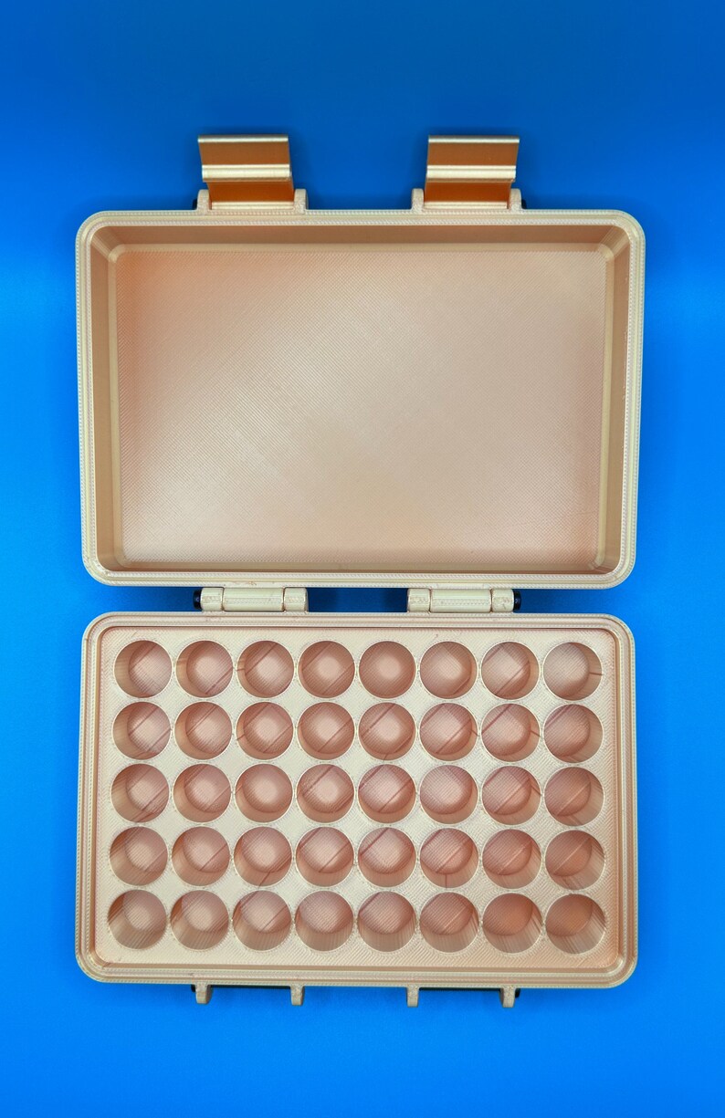 May include: A rectangular, metallic gold storage case with an open lid. The interior base features a grid of forty circular indentations arranged in five rows of eight, designed for holding small items. The case has two hinges and two latches.