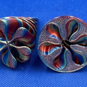 May include: Two 3D printed objects with a spiral, flower-like design. The objects feature a gradient of metallic red, blue, and gold colors with a textured surface finish. The shapes are hollow with curved, petal-like edges and a central opening.