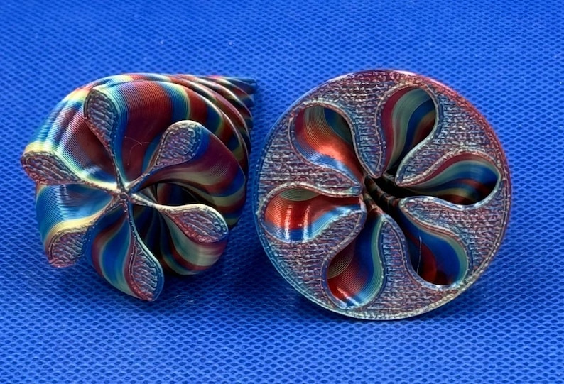 May include: Two 3D printed objects with a spiral, flower-like design. The objects feature a gradient of metallic red, blue, and gold colors with a textured surface finish. The shapes are hollow with curved, petal-like edges and a central opening.