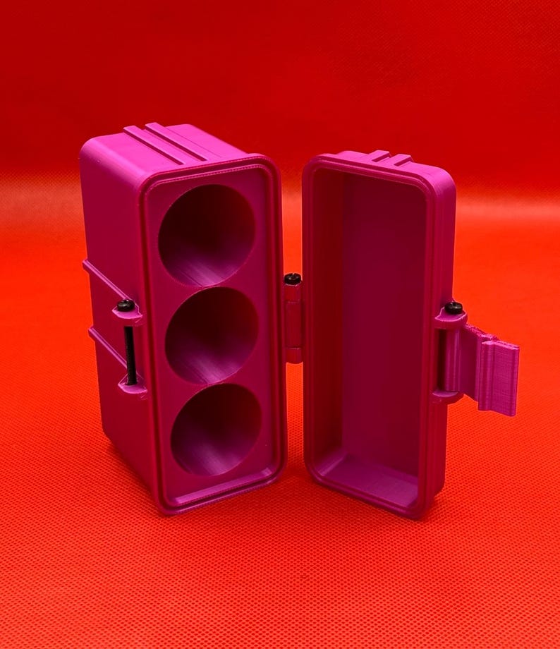 May include: A bright pink rectangular storage case with a hinged lid and a latch mechanism. The interior of the main body features three circular, vertically aligned compartments. The exterior has a textured, rigid surface design.
