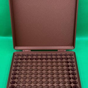 May include: A brown plastic case with a hinged lid and a foam interior with 100 round compartments. The case is designed to hold small items like pills, beads, or other small objects.