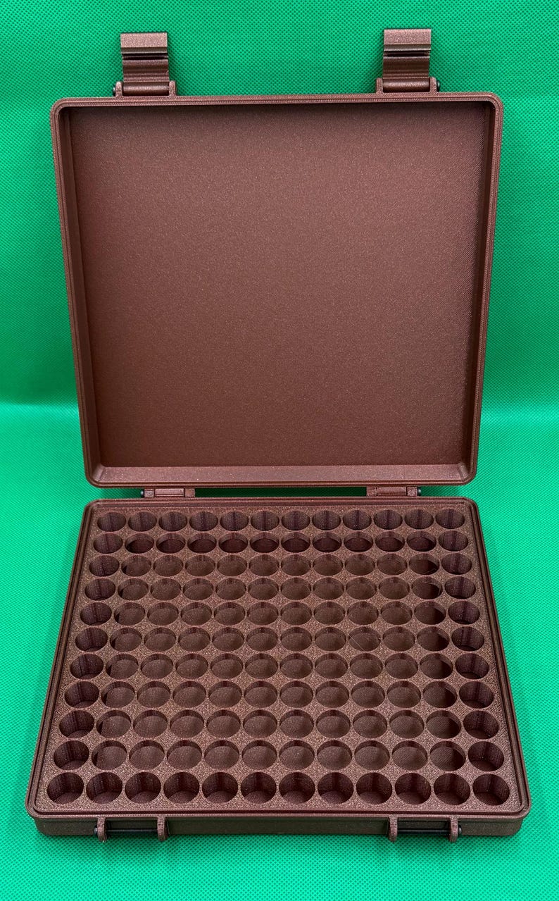 May include: A brown plastic case with a hinged lid and a foam interior with 100 round compartments. The case is designed to hold small items like pills, beads, or other small objects.