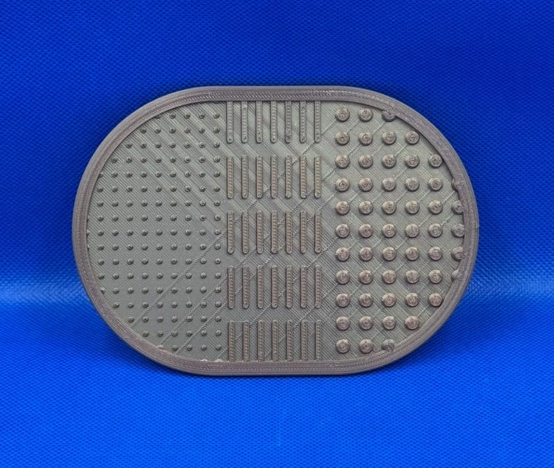 May include: An oval-shaped, metallic-brown makeup brush cleaning mat featuring a textured surface with three distinct sections: small raised dots on the left, vertical ridges in the center, and larger circular bumps on the right side.