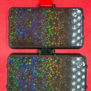 May include: A rectangular, hinged black plastic case featuring a holographic star pattern on both halves. The surface displays a gradient of iridescent, multi-colored star shapes against a dark background, with a red tab at the top.