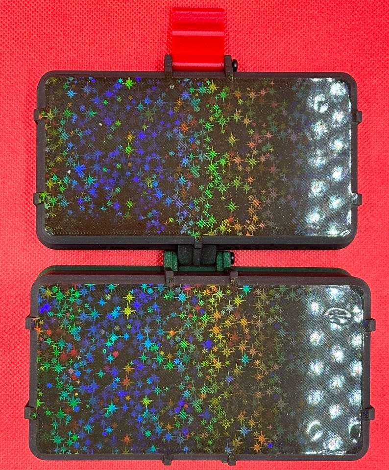 May include: A rectangular, hinged black plastic case featuring a holographic star pattern on both halves. The surface displays a gradient of iridescent, multi-colored star shapes against a dark background, with a red tab at the top.