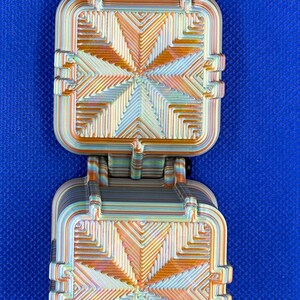 May include: A 3D printed fidget toy featuring two square, hinged components with a layered, iridescent finish. The surface displays a geometric, radiating pattern in shades of orange, gold, and teal, with a matching striped connector tab at the top.