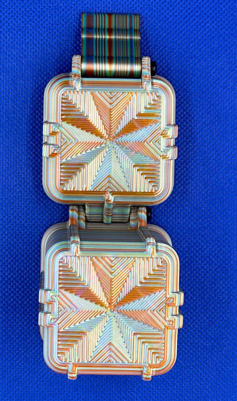 May include: A 3D printed fidget toy featuring two square, hinged components with a layered, iridescent finish. The surface displays a geometric, radiating pattern in shades of orange, gold, and teal, with a matching striped connector tab at the top.