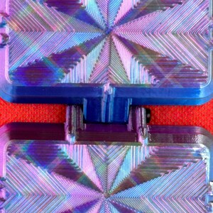 May include: A rectangular, hinged, two-part object featuring a vibrant, iridescent finish that shifts between shades of blue, purple, and pink. The surface displays a complex, geometric, starburst-like pattern of raised, linear ridges.