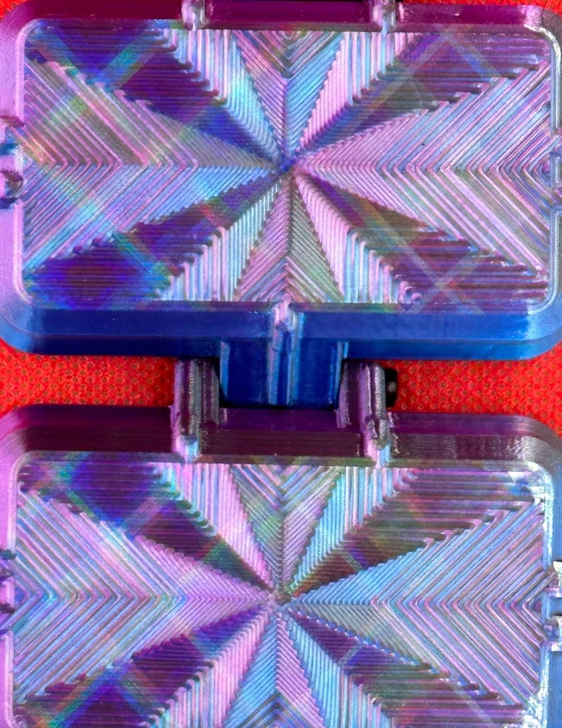 May include: A rectangular, hinged, two-part object featuring a vibrant, iridescent finish that shifts between shades of blue, purple, and pink. The surface displays a complex, geometric, starburst-like pattern of raised, linear ridges.