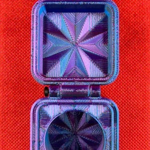 May include: A square, hinged container with a metallic, iridescent finish that shifts between blue and purple. The interior surfaces feature a textured, geometric starburst pattern, and the base includes a circular opening in the center.