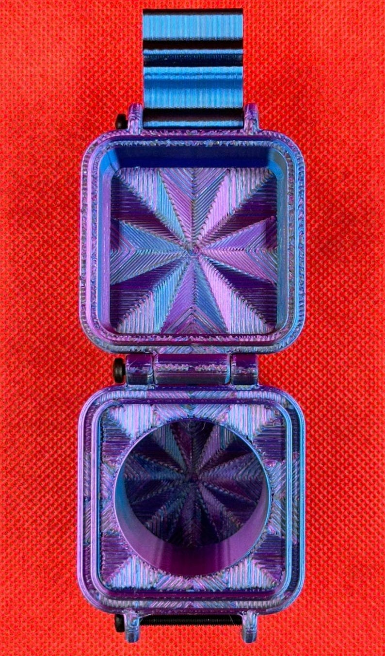 May include: A square, hinged container with a metallic, iridescent finish that shifts between blue and purple. The interior surfaces feature a textured, geometric starburst pattern, and the base includes a circular opening in the center.