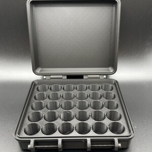 May include: A black, rectangular, open storage case featuring a grid of thirty circular compartments arranged in five rows of six. The case has a hinged lid with two top latches and a front locking mechanism, all constructed from a matte material.