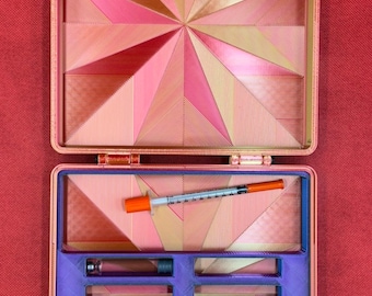 May include: A pink and gold geometric patterned case with a purple interior. The case has multiple compartments for storing small items, including a syringe and a pen.