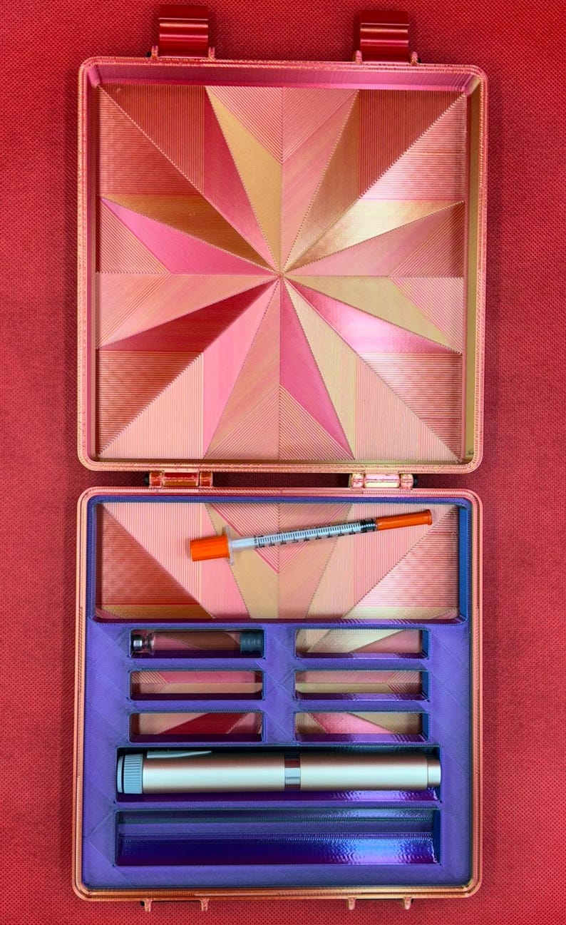 May include: A pink and gold geometric patterned case with a purple interior. The case has multiple compartments for storing small items, including a syringe and a pen.