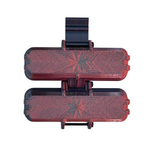 Puede incluir: Un dispositivo de sujeci&oacute;n de doble barra con dos componentes rectangulares dispuestos horizontalmente con un acabado texturizado en degradado rojo oscuro y negro. Cada componente muestra un patr&oacute;n de estrella y un soporte negro.