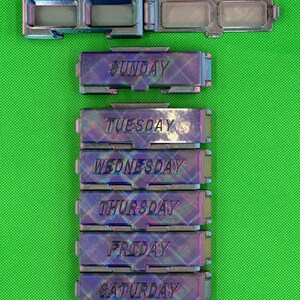May include: A set of six rectangular, iridescent, 3D-printed pill organizers with hinged lids. Each container is labeled with a day of the week: Sunday, Tuesday, Wednesday, Thursday, Friday, and Saturday. The surfaces feature a cross-hatch texture.