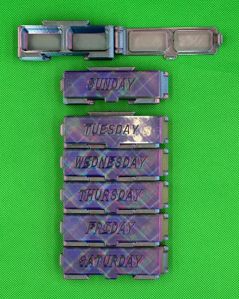 May include: A set of six rectangular, iridescent, 3D-printed pill organizers with hinged lids. Each container is labeled with a day of the week: Sunday, Tuesday, Wednesday, Thursday, Friday, and Saturday. The surfaces feature a cross-hatch texture.