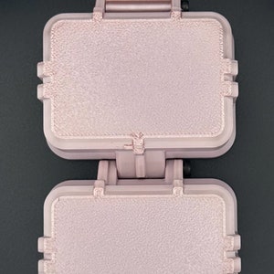 May include: A light pink, rectangular, dual-compartment hinged case with a textured surface finish. The device features a vertical orientation with a top mounting bracket and a black screw mechanism visible at the bottom hinge point.