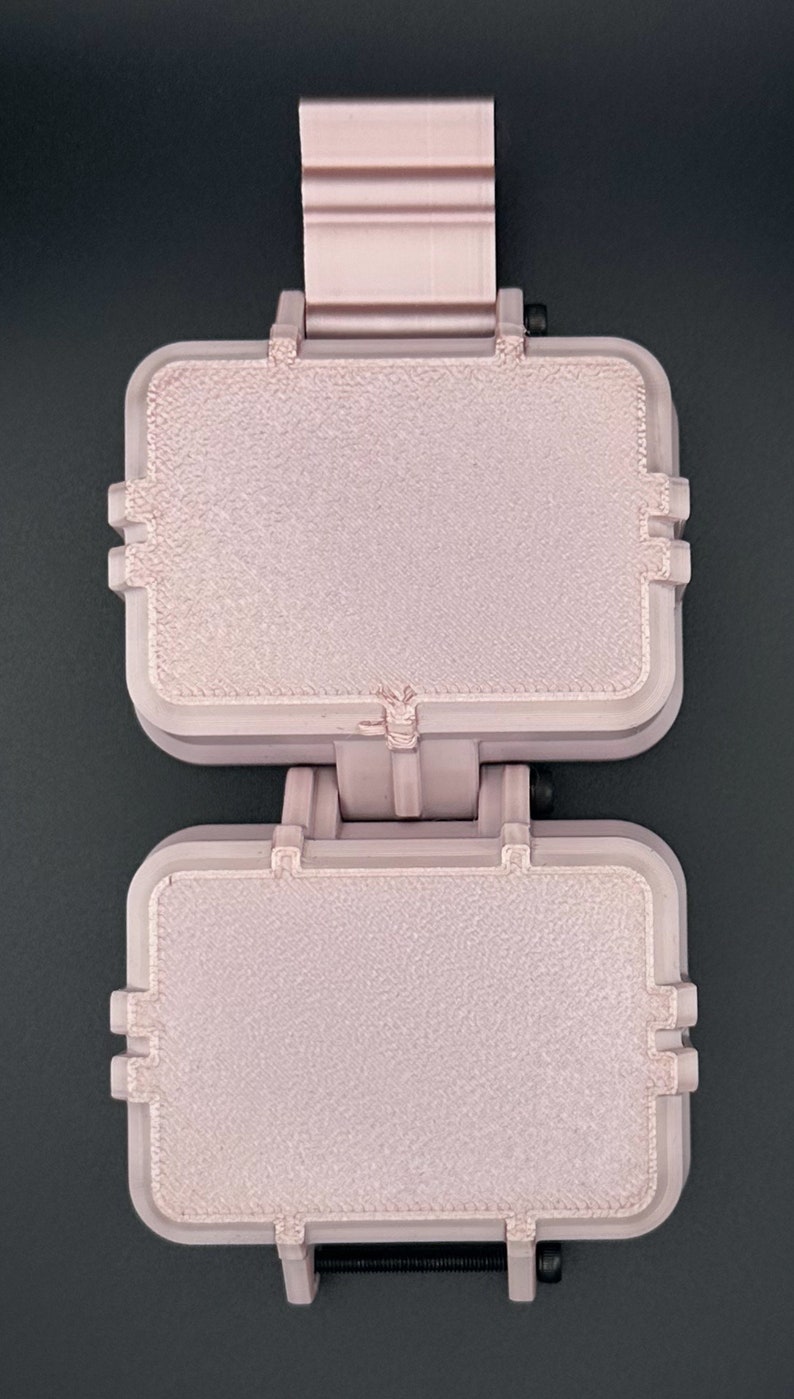May include: A light pink, rectangular, dual-compartment hinged case with a textured surface finish. The device features a vertical orientation with a top mounting bracket and a black screw mechanism visible at the bottom hinge point.