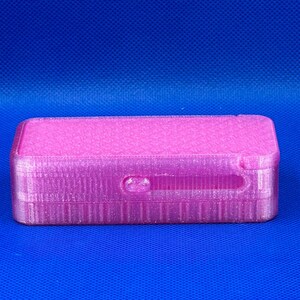 May include: A rectangular, 3D-printed pink storage box with a sliding lid mechanism. The surface features a subtle, repeating geometric grid pattern across the top and a smooth, layered texture along the sides of the container.
