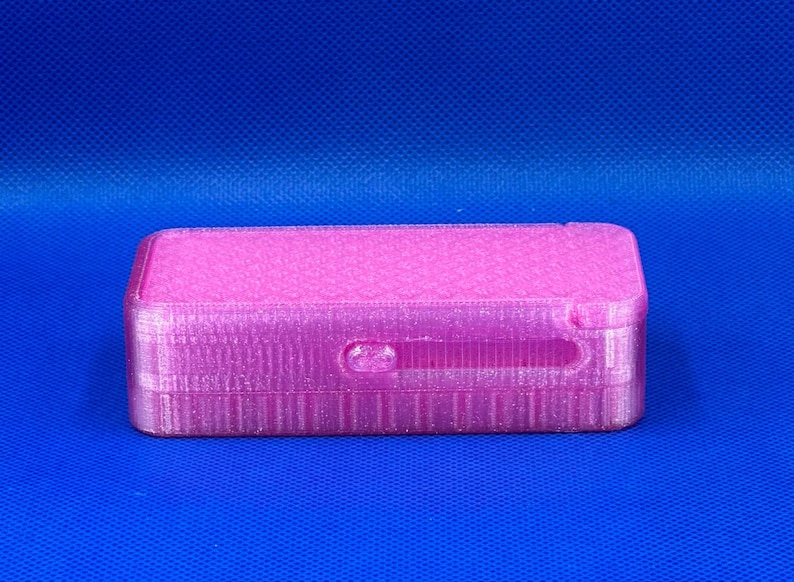 May include: A rectangular, 3D-printed pink storage box with a sliding lid mechanism. The surface features a subtle, repeating geometric grid pattern across the top and a smooth, layered texture along the sides of the container.