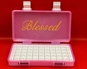 Könnte beinhalten: Ein rosafarbener, rechteckiger Aufbewahrungskoffer mit Klappdeckel, auf dem das Wort Blessed in gelber Schreibschrift steht. Das Innere enthält einen weißen Gittereinsatz mit mehreren runden Fächern zur Aufbewahrung kleiner Gegenstände, gesichert durch zwei silberfarbene Metallscharniere.