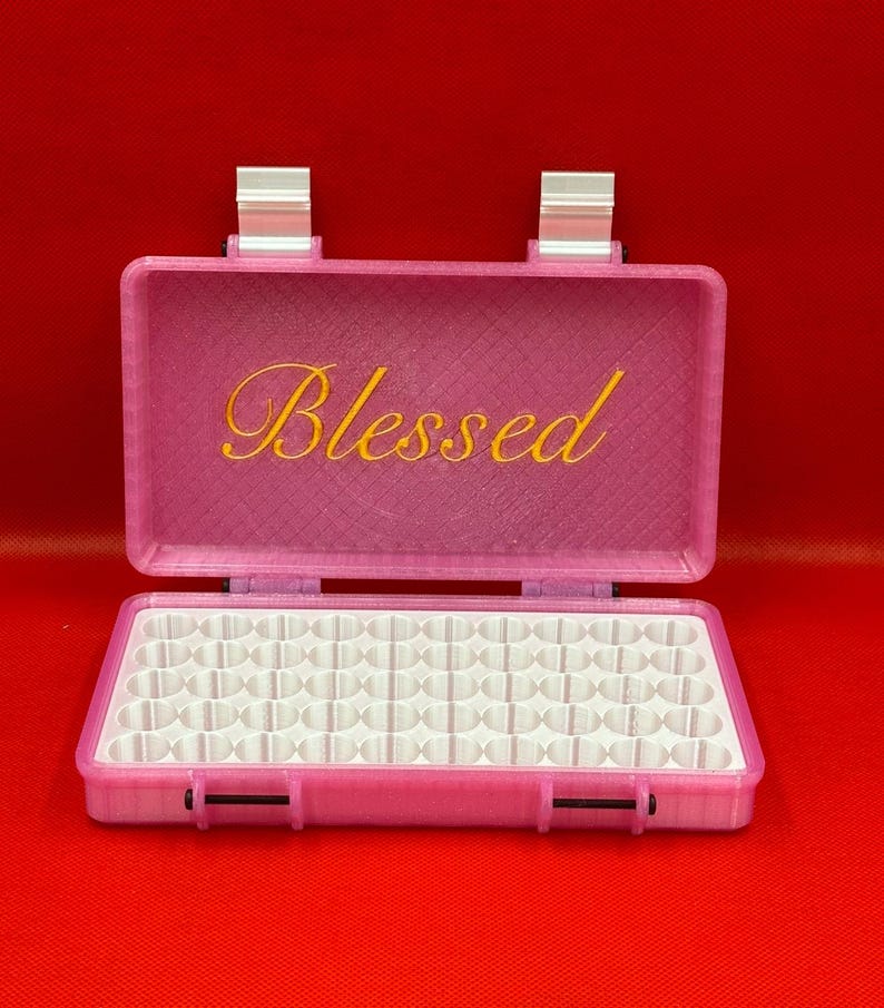 May include: A pink rectangular storage case with a hinged lid featuring the word Blessed in yellow script. The interior contains a white grid insert with multiple circular compartments for organising small items, secured by two silver-coloured metal hinges.