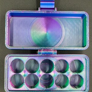 May include: A rectangular, hinged storage case with a vibrant, iridescent gradient finish shifting between blue, purple, and green. The top section is an open compartment, while the bottom section features ten circular cutouts for storage.