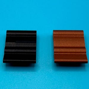 May include: Two rectangular clips with a wavy, grooved surface profile. One clip is solid black and the other is a metallic copper color. Both items feature a consistent, repeating horizontal ridge pattern across their top surfaces.