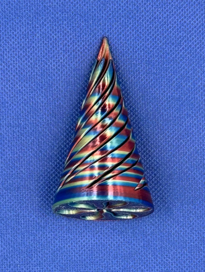 May include: A conical object featuring a metallic finish with a swirling, iridescent pattern of red, blue, and gold colors. The surface has deep, diagonal grooves that spiral toward the pointed top, and the base has a star-shaped cutout.