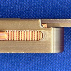 May include: A rectangular metallic object with a brushed bronze finish featuring a sliding mechanism. A copper-coloured textured thumb grip is set into a recessed slot on the side, allowing for manual adjustment of the sliding component.