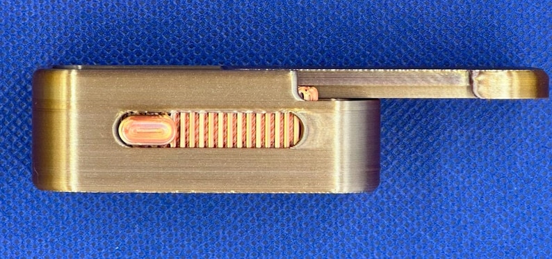 May include: A rectangular metallic object with a brushed bronze finish featuring a sliding mechanism. A copper-coloured textured thumb grip is set into a recessed slot on the side, allowing for manual adjustment of the sliding component.