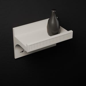 Può includere: Una mensola da parete bianca con bordo anteriore scanalato e supporto curvo. Un vaso nero opaco con collo stretto e corpo arrotondato poggia sulla superficie piana della mensola. La mensola presenta un piccolo intaglio sul lato.