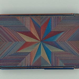 May include: Rectangular case with a starburst design. The case features a geometric pattern in shades of red, blue, and tan. The case has a metallic finish and a hinged lid.