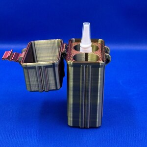 May include: A rectangular, metallic-toned 3D printed storage case with a hinged lid. The case features two circular interior compartments, one of which holds a small white plastic spray bottle with a pointed nozzle tip.