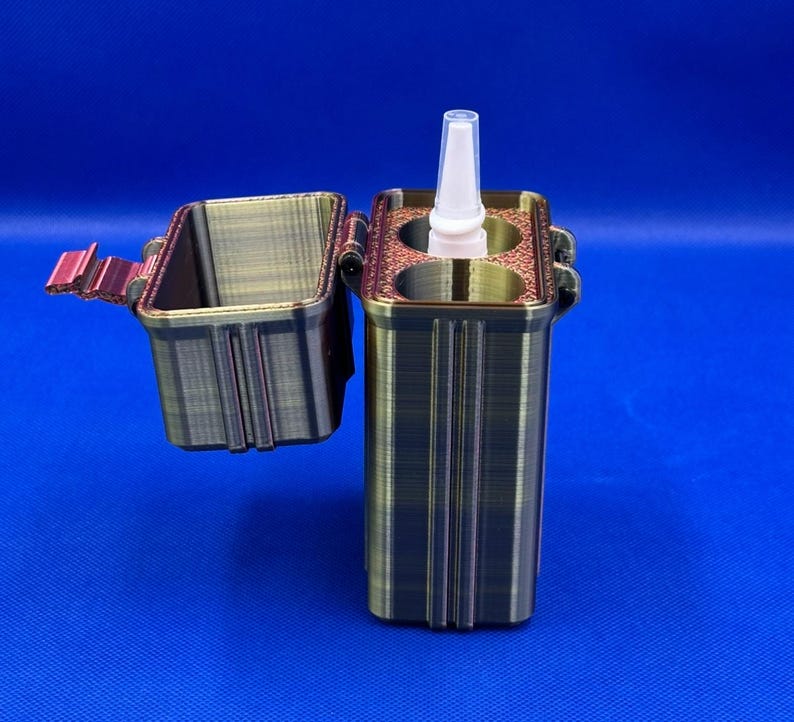 May include: A rectangular, metallic-toned 3D printed storage case with a hinged lid. The case features two circular interior compartments, one of which holds a small white plastic spray bottle with a pointed nozzle tip.