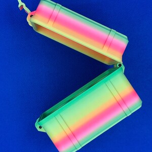 May include: A rectangular storage container with an open hinged lid, featuring a vibrant gradient color pattern that transitions through shades of pink, orange, yellow, and green. The box has a textured exterior with vertical ridges.