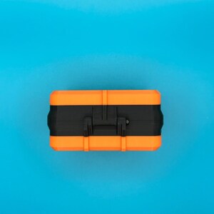 May include: A rectangular storage case featuring a vibrant orange top and bottom section separated by a wide black centre band. The case includes a central black handle mechanism integrated into the middle section, designed for secure closure.