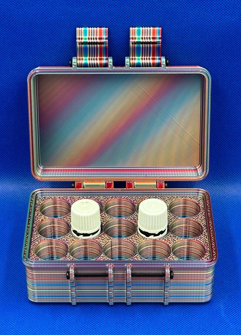 May include: A rectangular storage case with a hinged lid, featuring a multi-colored gradient pattern of horizontal and diagonal lines. The interior contains twelve circular compartments, with two white cylindrical caps visible inside.