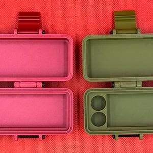 以下が含まれることがあります： 鮮やかなピンクとオリーブグリーンの長方形のオープン収納ケース2つ。各ケースにはヒンジ付きの蓋、2つの留め具、そして小さなアイテムを収納するための左側に2つの円形のくぼみがある内部コンパートメントが備わっています。